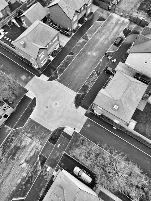 An aerial black-and-white photograph of a residential street showing several houses with pitched roofs and small front gardens. The street features a central intersection with crosswalks and paved pathways connecting to each property. parked cars are visible along the curbs, including one positioned on the pavement near a tree with bare branches. The area appears calm and well-maintained, with no visible damage to the pavement or surrounding environment. The image captures a scene related to home relocation or moving logistics, with the arrangement of houses, roadways, and parked vehicles illustrating the typical setting where professional removals services such as those by Man with Van Ickenham may operate during house removals or furniture transport in the Ickenham area.