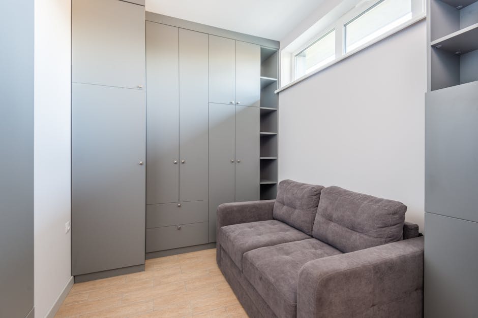 The image shows a compact interior room with light wood flooring and white painted walls. Against one wall, there is a large, modern grey storage unit with multiple cabinet doors and open shelving sections, some containing small items. On an adjacent wall near the window, which provides natural light, there is a grey fabric sofa with a simple, contemporary design, seating two or three people. The window is narrow and horizontal, positioned high on the wall, allowing daylight to illuminate the space. The room appears tidy and well-organized, potentially part of a home undergoing furniture arrangements or preparing for moving. This setting may be related to house removals or packing and moving services handled by Man with Van Ickenham, who specialise in furniture transport and home relocation logistics, as indicated on the webpage for bulky item disposal in Ickenham.