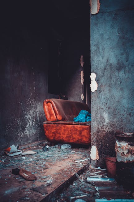 A partially visible red upholstered sofa with a curved backrest and armrest, situated in a dusty, dilapidated interior with damaged black and grey walls showing chipped paint and exposed brickwork. The sofa has a crumpled blue cloth on it, and the surrounding floor is covered in debris, including scattered bricks, dust, and rubbish. The room appears neglected, with an uneven floor and a small section of a doorway visible on the left side. This scene reflects a space undergoing clearance or preparation for a home relocation, related to furniture transport and packing and moving services. The image is in natural light, capturing the current state of disuse, aligning with themes of house removals and bulky item disposal in Ickenham, as operated by Man with Van Ickenham.