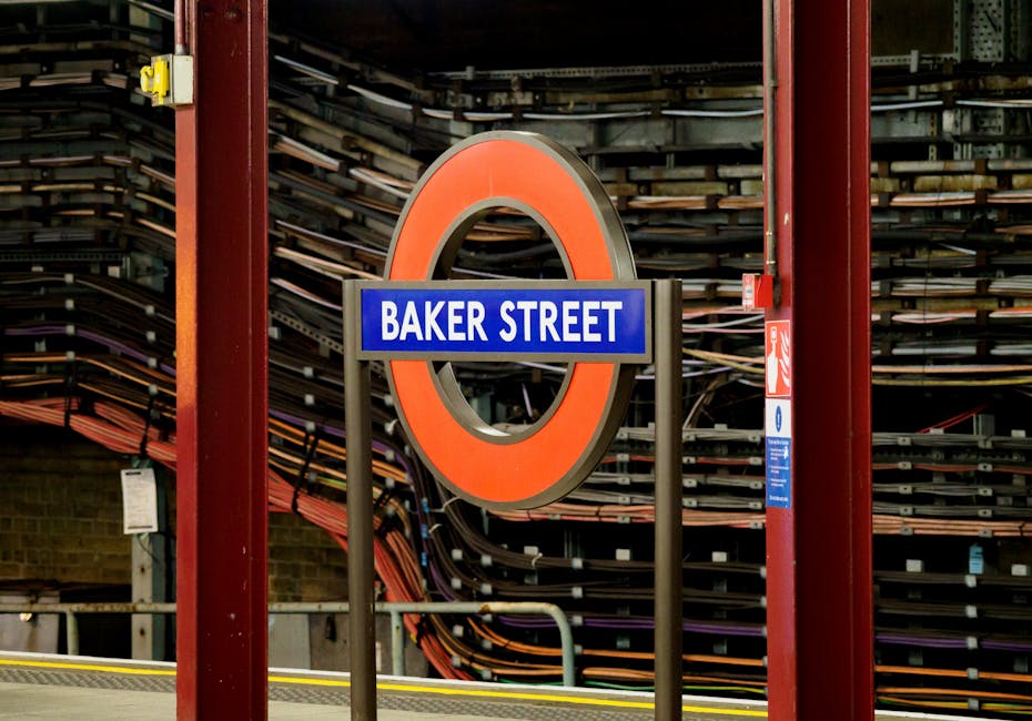 A close-up view of a London Underground station sign displaying 'Baker Street' in white text on a blue background, mounted on a metal post. Behind the sign, there are large, stacked railway lines or track panels in shades of brown, black, and grey, neatly organized against a brick wall. The station structure features red metal supports around the sign and adjacent signage, with the platform surface visible at the bottom. The setting appears to be inside the station, illuminated by artificial lighting, reflecting a typical urban environment associated with house removals and furniture transport for homes near London Tube stations. This image is related to transportation logistics involved in home relocation, particularly the route and infrastructure used by companies like Man with Van Ickenham to facilitate efficient moving services.