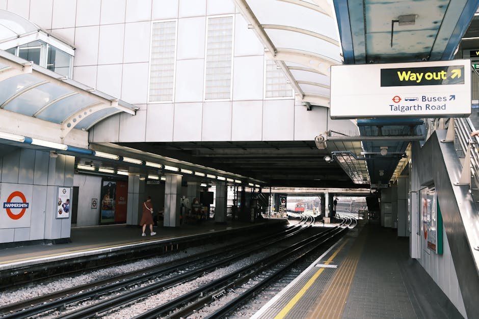 The entrance to Hammersmith underground station in London features an outdoor area with train tracks running through the station. The platform has a tiled surface and is covered by a modern, curved transparent canopy supported by metal beams. Several passengers are visible on the platform, some walking and others waiting. On the right side, a staircase with metal railings leads up to the station entrance, which is marked by signs including a yellow 'Way out' sign with an arrow and directional signage to buses on Talgarth Road. The station walls are tiled in white and decorated with London Underground roundel logos. The overall scene illustrates the typical setting of a busy urban underground station in the context of home relocation or furniture transport, with the environment prepared for efficient passenger and baggage movement, aligning with house removals and moving services like those offered by Man with Van Ickenham.
