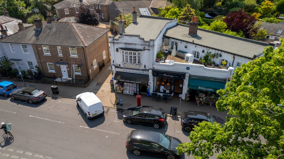 An aerial view of a street scene in Ickenham, showing a row of small shops with distinct architectural features, including a white building with an ornate facade and a curved roofline, and a darker building with a green awning. In front of the shops, three cars are parked along the pavement, with a white van positioned near the curb, likely involved in a home relocation or furniture transport process. The sidewalk is partly shaded by leafy green trees, and a few pedestrians are nearby. The background features residential houses with gardens and trees, indicating a suburban setting. This image captures the urban environment typical of Ickenham, illustrating the loading process for house removals, with packaging materials or furniture possibly being loaded or unloaded from the vehicles while Man with Van Ickenham manages the move.