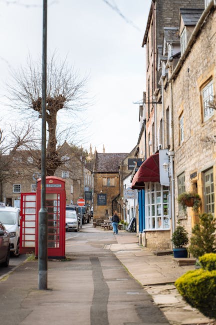 A quiet residential street in Ickenham with traditional brick houses featuring pitched roofs and multiple chimneys, some with dormer windows. The pavement runs alongside the front of the houses, with a black wrought iron lamppost positioned near the corner on the left side. A large green tree stands to the right, partially obscuring a white house with decorative architectural details at the entrance. In the middle distance, a dark grey van is parked on the road, which gently curves to the right. The scene appears to be during daylight hours with overcast sky, indicating a typical setting for home relocation or furniture transport activities, as part of professional removals services like those offered by Man with Van Ickenham. The overall environment suggests a peaceful, well-maintained neighbourhood suitable for house removals or moving services, with clear visibility of the street and nearby housing.