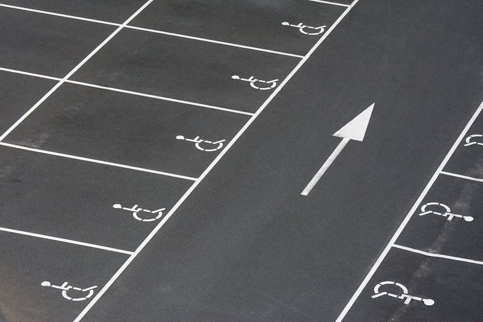 Photograph of a section of a paved parking area with marked spaces designated for disabled access, indicated by white wheelchair symbols on the dark asphalt. An arrow painted on the ground points forward, guiding vehicle movement within the lot. The parking bays are outlined with white lines, with some spaces partially visible on both sides of the image. The surface appears clean, and the markings are clearly visible. This scene demonstrates typical parking lot organization relevant to moving and relocation logistics, as used by services such as Man with Van Ickenham when loading or unloading furniture and boxes during home relocation or house removals in Ickenham. The image captures the outdoor environment, emphasizing parking arrangements pertinent to logistical planning for household moves, ensuring access and efficient vehicle maneuvering.
