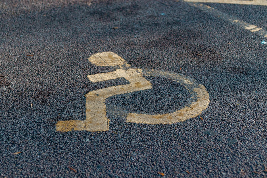 Close-up view of a parking lot with a dark asphalt surface, showing faded white line markings and a large, worn bicycle symbol within a marked parking space. The asphalt texture is rough, with some small debris scattered across the surface. The scene is illuminated by natural daylight, with no vehicles or objects visible beyond the parking markings. This setting is typical of residential or street parking areas used during home relocation or furniture transport processes. Such parking spaces are often utilized by professional removals services, like Man with Van Ickenham, when loading or unloading items for moves such as those described in the Swakeleys Park moves advice page regarding access, parking, and lift considerations.