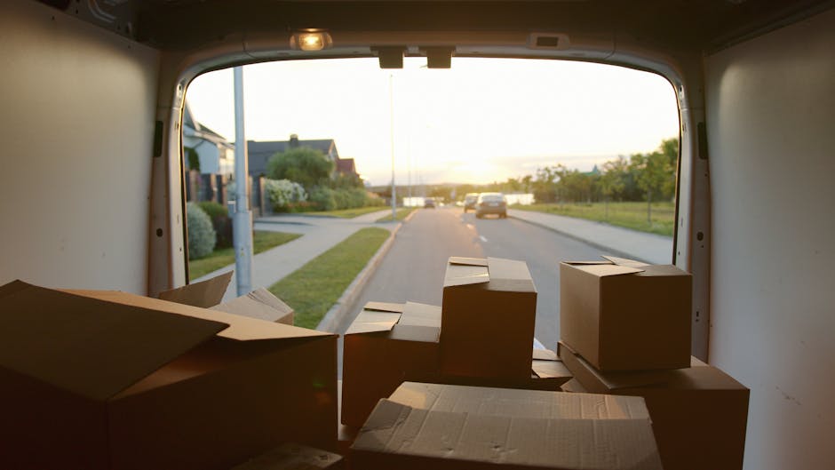 View from inside a moving van parked on a residential street during sunset, showing several packed cardboard boxes of varying sizes placed on the floor near the open rear door. The boxes are sealed with tape, and some are stacked while others lie flat, ready for home relocation or furniture transport. Visible in the background are parked cars, a sidewalk, and houses with front gardens, under a partly cloudy sky with warm lighting. The interior of the van is illuminated by natural light, highlighting the boxes prepared for loading and transport, as Man with Van Ickenham continues with packing and moving services for local house removals.
