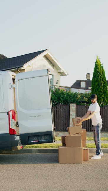 A young man wearing a white t-shirt and jeans is standing on a suburban street during daytime, preparing cardboard boxes for loading into a white transit van parked next to a residential property. He is holding a box in his hands, with several other cardboard boxes placed on the pavement nearby, some stacked and others ready for transport. The van's rear doors are open, revealing a partially visible interior with a plastic-wrapped item and other packing materials, indicating a home relocation or furniture transport process. The background features residential houses, a wooden fence, well-maintained green bushes, and a tall coniferous tree, under a clear sky. Man with Van Ickenham’s service involves packing and loading activities aligned with house removals and moving logistics, as seen in this loading process scene.