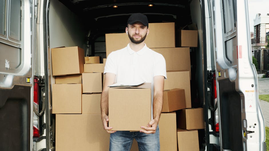 View from inside a moving van parked on a residential street during sunset, showing several packed cardboard boxes of varying sizes placed on the floor near the open rear door. The boxes are sealed with tape, and some are stacked while others lie flat, ready for home relocation or furniture transport. Visible in the background are parked cars, a sidewalk, and houses with front gardens, under a partly cloudy sky with warm lighting. The interior of the van is illuminated by natural light, highlighting the boxes prepared for loading and transport, as Man with Van Ickenham continues with packing and moving services for local house removals.