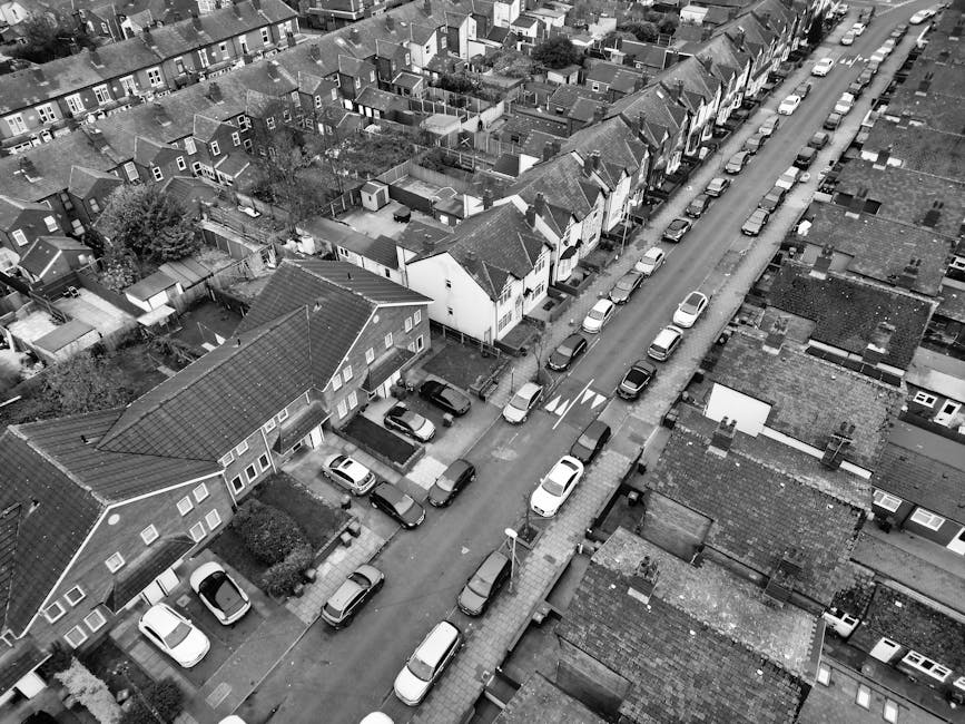 An aerial view in black and white showing a residential street lined with parked cars on both sides. Behind the parked vehicles, rows of terraced houses with pitched roofs extend along the narrow road, some with small front gardens or driveways. A lorry from Man with Van Ickenham is partially visible on the right side, positioned on the pavement next to a house with an open doorway, indicating the loading process for house moves. The scene captures a typical suburban neighbourhood with closely packed houses and parked vehicles, illustrating the logistical considerations of house removals and furniture transport on narrow roads with limited parking space, relevant to the UB10 area and Ickenham property relocations.