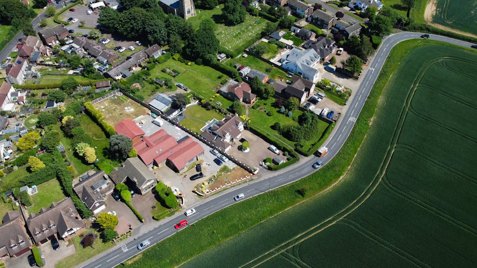 An aerial view of a residential area showing a narrow street with several parked cars and moving vehicles, bordered by houses with well-maintained gardens and fences. Some houses have driveways where moving boxes, cardboard cartons, and furniture covered with protective blankets are being loaded onto a large white van parked on the side of the road. Visible packing materials such as plastic wrap and wooden furniture slats are positioned near the loading process, with a person actively carrying a box towards the van, indicating a home relocation or furniture transport operation. The surrounding environment includes green fields and curved roadways, highlighting the logistical challenges of house moves in narrow roads in Ickenham, relevant to the services offered by Man with Van Ickenham in their removals category.