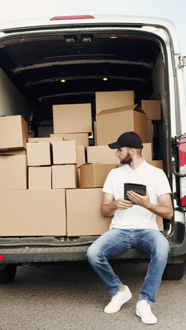 A man wearing a white T-shirt, blue jeans, white sneakers, and a black cap is sitting on the pavement beside the open rear doors of a white van, which is filled with numerous packed cardboard boxes of various sizes, some stacked on top of each other and others placed side by side. The boxes are sealed with packing tape, and a few are partly covered with fabric or plastic wrapping, indicating a home relocation process. Inside the van, the interior is dark, and the boxes are arranged in an organized manner to facilitate loading and transportation. The man is holding a tablet device, possibly reviewing a moving checklist or inventory. The scene takes place outdoors with a neutral background and natural lighting, illustrating the logistics involved in furniture transport and packing during a professional removals service, as provided by Man with Van Ickenham, supporting efficient house removals and relocation services.