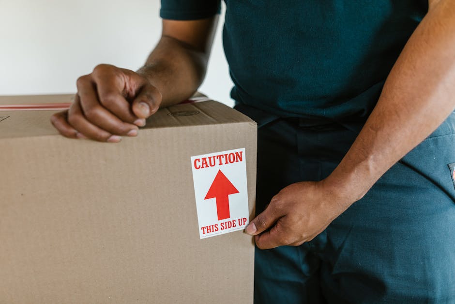 A person wearing a dark blue t-shirt is holding a large cardboard box with both hands, positioning it on a flat surface. The box features a red and white caution label with an upward-pointing arrow and the text 'Caution - This Side Up.' Behind the person, part of a moving van or vehicle is visible, indicating an active loading process for home relocation or packing and moving services. The setting appears indoors, with natural lighting illuminating the scene, and additional packing materials or smaller boxes may be present in the background, supporting efficient furniture transport and moving logistics handled by Man with Van Ickenham.
